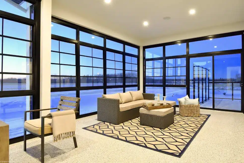 A cozy, modern sunroom interior with black framing and a large sectional sofa, looking out over a snow-covered backyard.